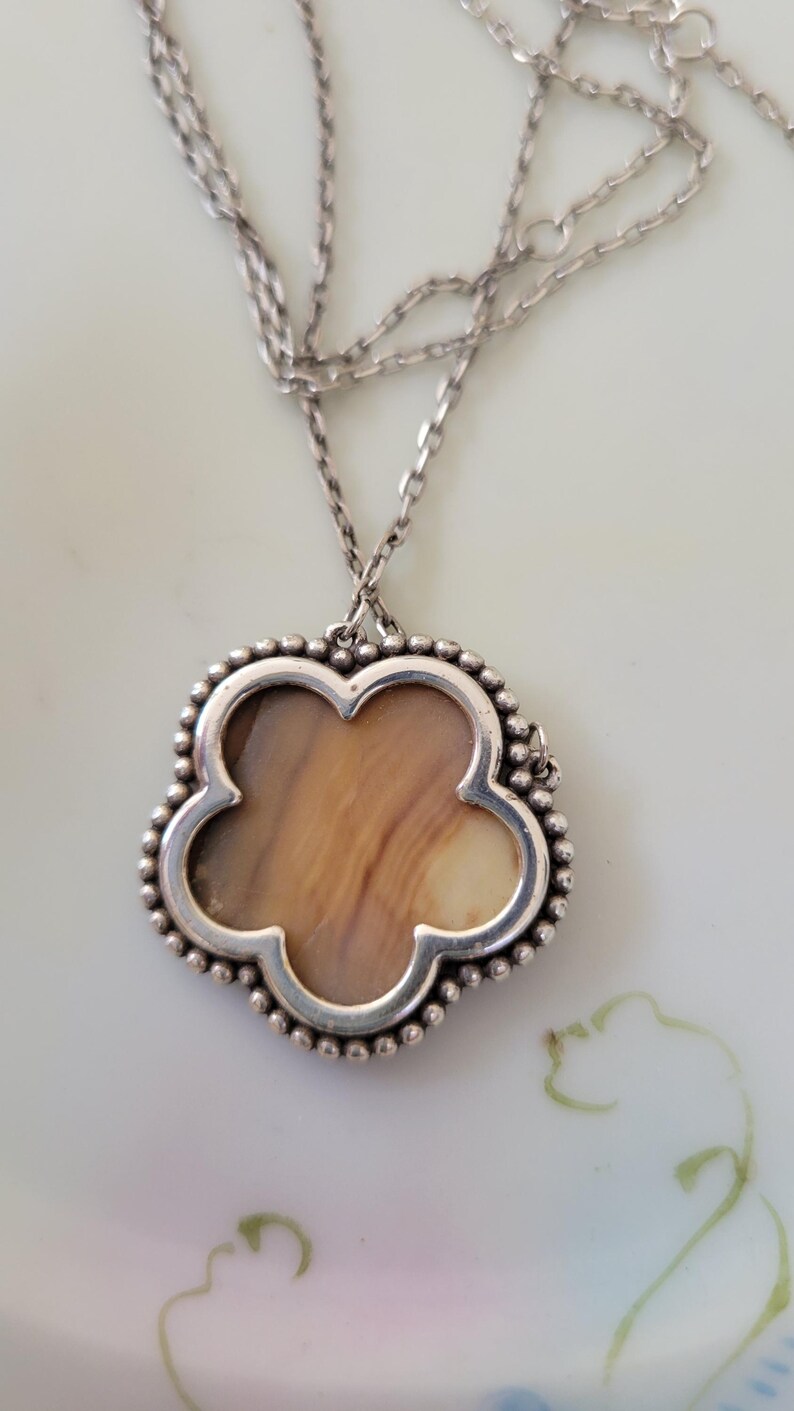 May include: A silver necklace with a clover-shaped pendant. The pendant has a light brown, patterned center and a beaded silver border. The chain is also silver and has a delicate design. The necklace is displayed on a white surface.