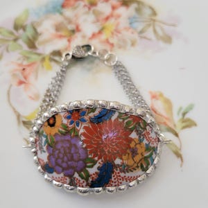 May include: A silver bracelet featuring an oval centerpiece with a floral design in shades of red, purple, yellow, and blue. The bracelet has a double chain and a decorative border around the centerpiece. The clasp is heart-shaped.
