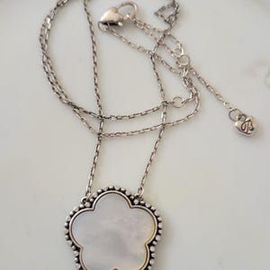 May include: Silver-toned necklace with a flower-shaped pendant. The pendant has a mother-of-pearl center and a beaded border. The necklace chain is delicate, and the clasp has a heart-shaped charm.