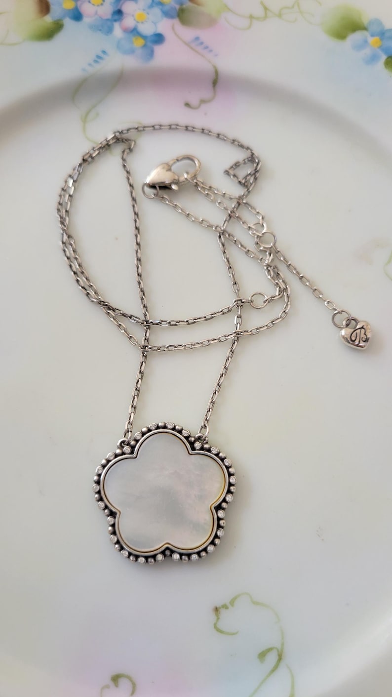 May include: Silver-toned necklace with a flower-shaped pendant. The pendant has a mother-of-pearl center and a beaded border. The necklace chain is delicate, and the clasp has a heart-shaped charm.
