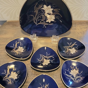 May include: A set of seven triangular ceramic dishes featuring a deep navy blue glaze with gold-toned lily floral illustrations. The collection includes one large serving dish and six smaller matching plates, all finished with a gold-toned rim.