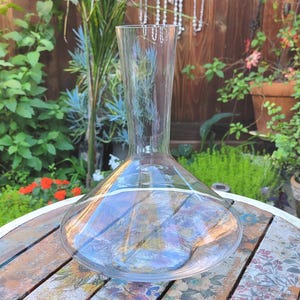 May include: A clear glass wine decanter with a wide base and a tall, slender neck. The decanter sits on a wooden table with a floral pattern. The background features lush greenery and flowers.