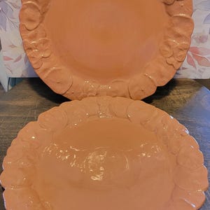 May include: Two round ceramic plates in a solid peach color. Each plate features a scalloped edge decorated with a repeating embossed floral pattern of pansies or similar blossoms around the entire rim, creating a textured border.