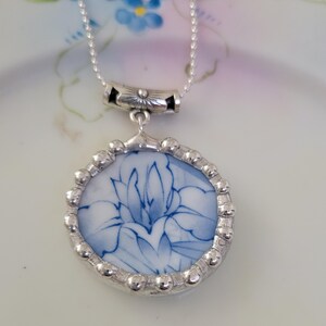 May include: A circular pendant necklace featuring a piece of white ceramic with a blue floral pattern, encased in a silver-toned metal setting with a beaded edge. The pendant hangs from a silver-toned chain attached to a decorative metal bail.