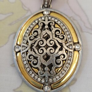 Puede incluir: Collar con colgante ovalado con un dise&ntilde;o plateado y dorado. El colgante presenta un intrincado dise&ntilde;o de filigrana en el centro, rodeado por un borde dorado y peque&ntilde;os cristales transparentes. El collar tiene una cadena plateada.