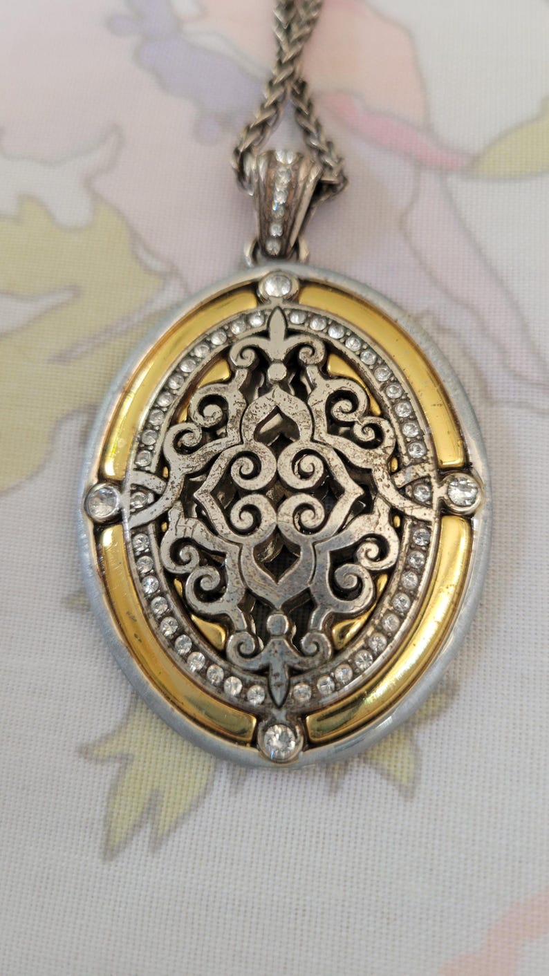 Puede incluir: Collar con colgante ovalado con un dise&ntilde;o plateado y dorado. El colgante presenta un intrincado dise&ntilde;o de filigrana en el centro, rodeado por un borde dorado y peque&ntilde;os cristales transparentes. El collar tiene una cadena plateada.