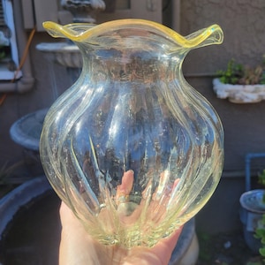 May include: A clear glass vase with a ruffled, yellow-tinted rim. The vase has a bulbous shape with vertical, textured ridges. The light reflects off the glass, creating a shimmering effect. The vase is held in a person's hand.