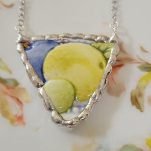 May include: A triangular pendant necklace featuring a ceramic shard with a yellow lemon and green leaf design on a blue background, encased in a textured silver-toned metal border and suspended from a delicate silver-toned chain.