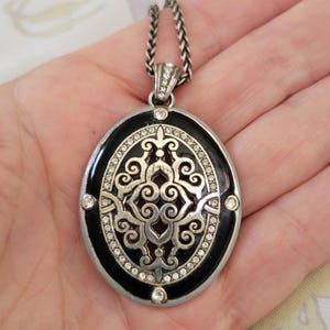 Puede incluir: Collar con colgante plateado con un colgante ovalado de esmalte negro. El colgante presenta un dise&ntilde;o decorativo de filigrana plateada, acentuado con cristales transparentes. La cadena del collar es trenzada.
