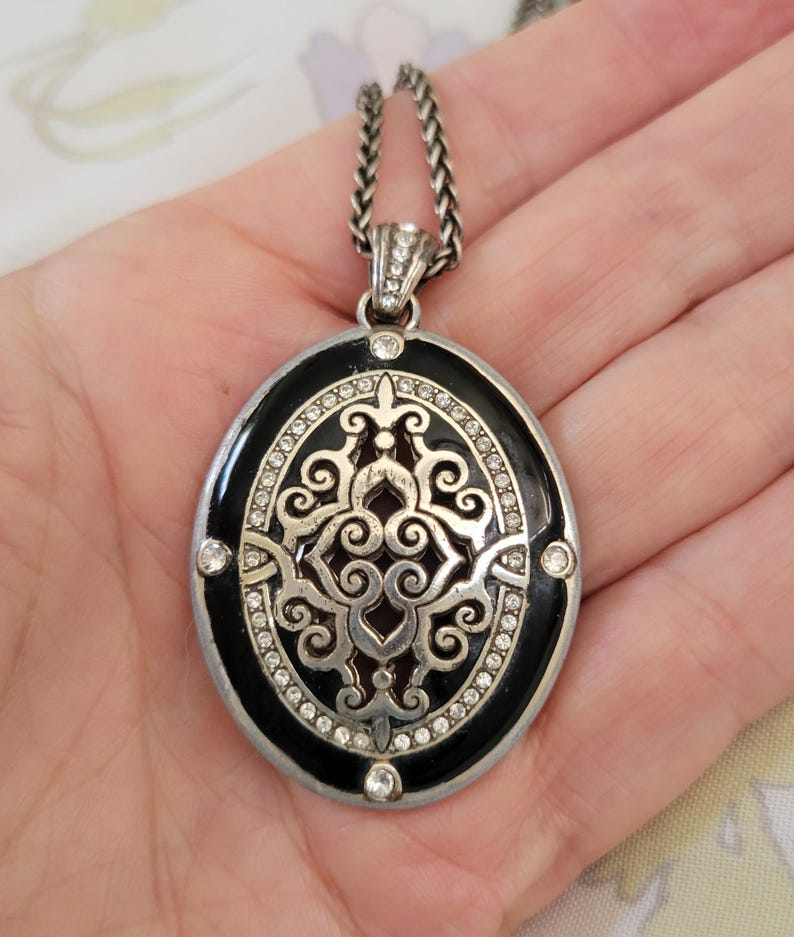 Puede incluir: Collar con colgante plateado con un colgante ovalado de esmalte negro. El colgante presenta un dise&ntilde;o decorativo de filigrana plateada, acentuado con cristales transparentes. La cadena del collar es trenzada.