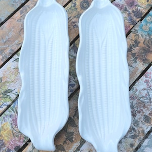 May include: Two white ceramic corn-shaped serving dishes. The dishes have a detailed texture mimicking corn kernels and leaves. The dishes are placed on a wooden surface with a floral pattern.