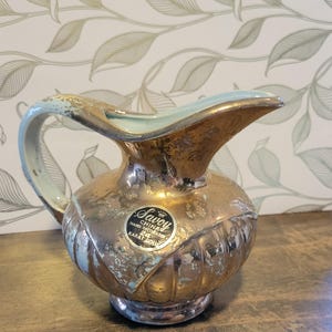 May include: A Savoy China pitcher with a metallic gold finish and a light blue interior. The pitcher features a rounded body with vertical fluting and a circular sticker that reads Savoy China Hand Decorated 24 Karat Gold.