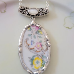 May include: Silver-toned necklace featuring an oval pendant with a floral design. The pendant is made from a piece of white porcelain with a colorful floral pattern. The necklace has a decorative bar and chain.