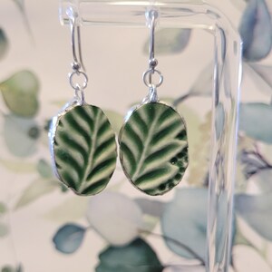 May include: A pair of dangle earrings with oval-shaped green and white leaf designs. The earrings have silver-colored hooks and are displayed on a clear stand. The leaf pattern is detailed with dark green veins.