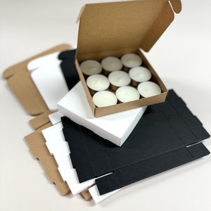 May include: A stack of cardboard boxes in brown, white, and black. The top box is open and contains a set of 10 white tea light candles.