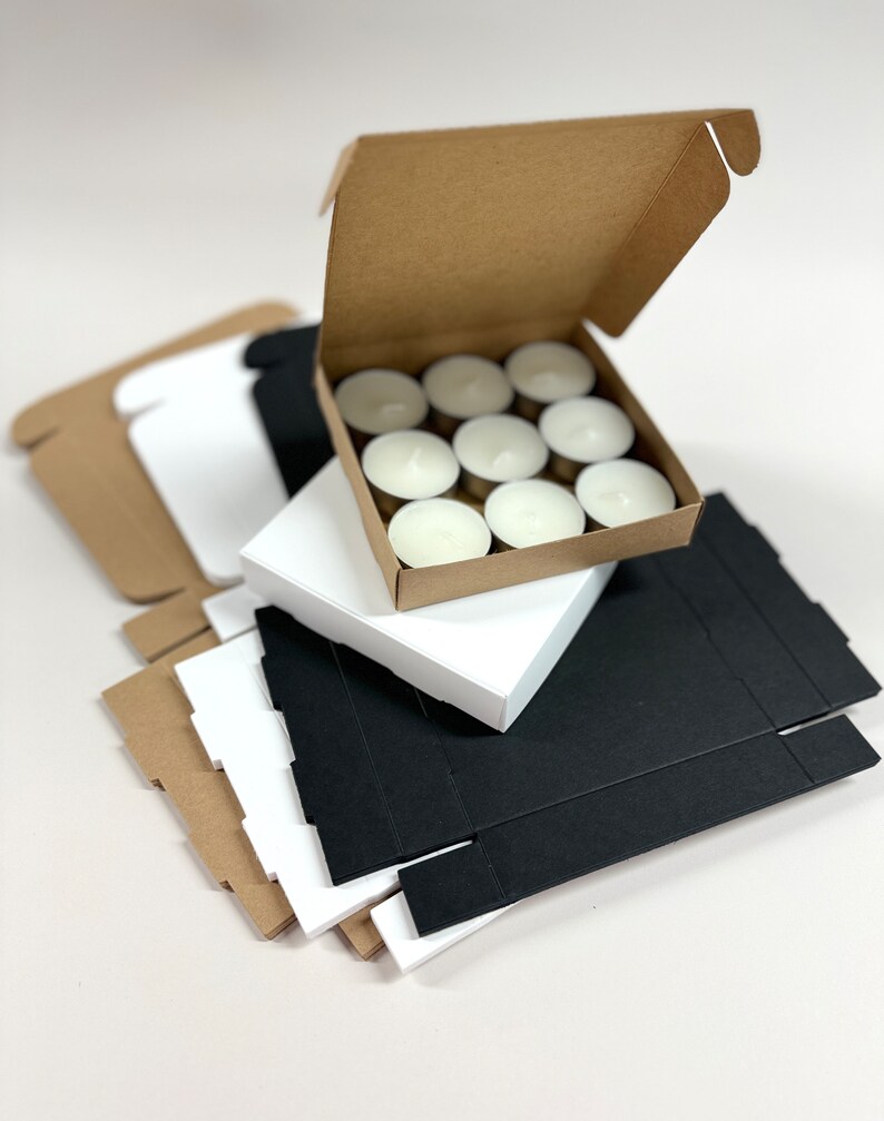 May include: A stack of cardboard boxes in brown, white, and black. The top box is open and contains a set of 10 white tea light candles.