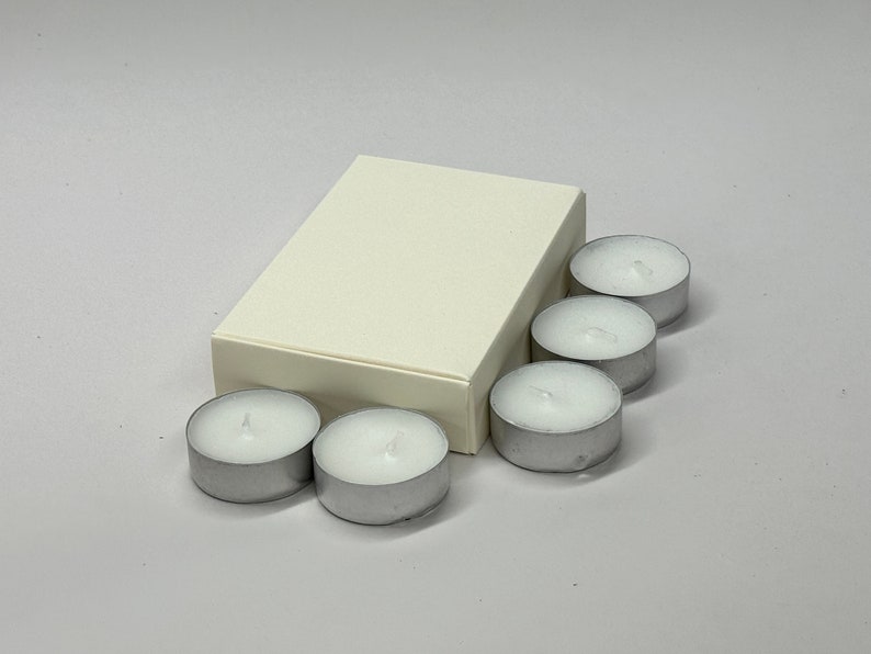 May include: A white box with six white tea light candles. The candles are in a silver holder.
