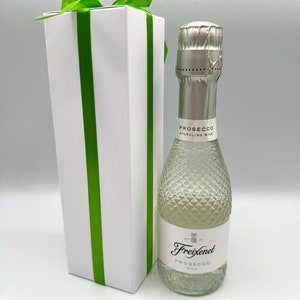 May include: A white gift box with a green ribbon tied around it. The box is next to a small bottle of Freixenet Prosecco sparkling wine.