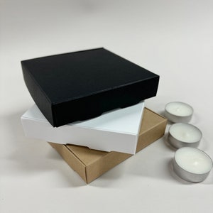 May include: Three square boxes stacked on top of each other. The top box is black, the middle box is white, and the bottom box is brown. Three small white tea lights are in the background.