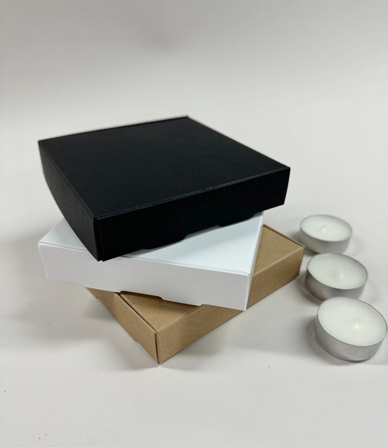 May include: Three square boxes stacked on top of each other. The top box is black, the middle box is white, and the bottom box is brown. Three small white tea lights are in the background.