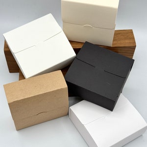 May include: Five small gift boxes in different colours. The boxes are made of cardboard and have a simple design. The colours are white, black, brown, and cream.