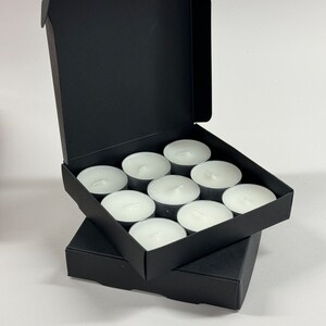 May include: A black box with a lid open, revealing a set of 10 white tea lights. The tea lights are arranged in a grid pattern inside the box.