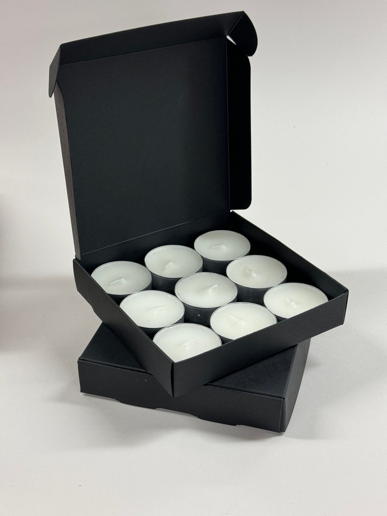 May include: A black box with a lid open, revealing a set of 10 white tea lights. The tea lights are arranged in a grid pattern inside the box.