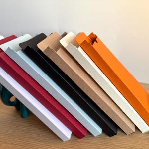 May include: A set of ten cardboard boxes in various colours, including white, red, light blue, black, brown, and orange. The boxes are stacked on top of each other, with the orange box on top. The boxes are all the same size and shape, and they have a flap on one side that can be folded down to close the box.