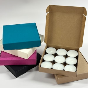 May include: A set of 10 white tea lights in a cardboard box. The box is open and the tea lights are visible. There are four empty boxes stacked on top of the box with tea lights. The boxes are teal, white, pink, and black.