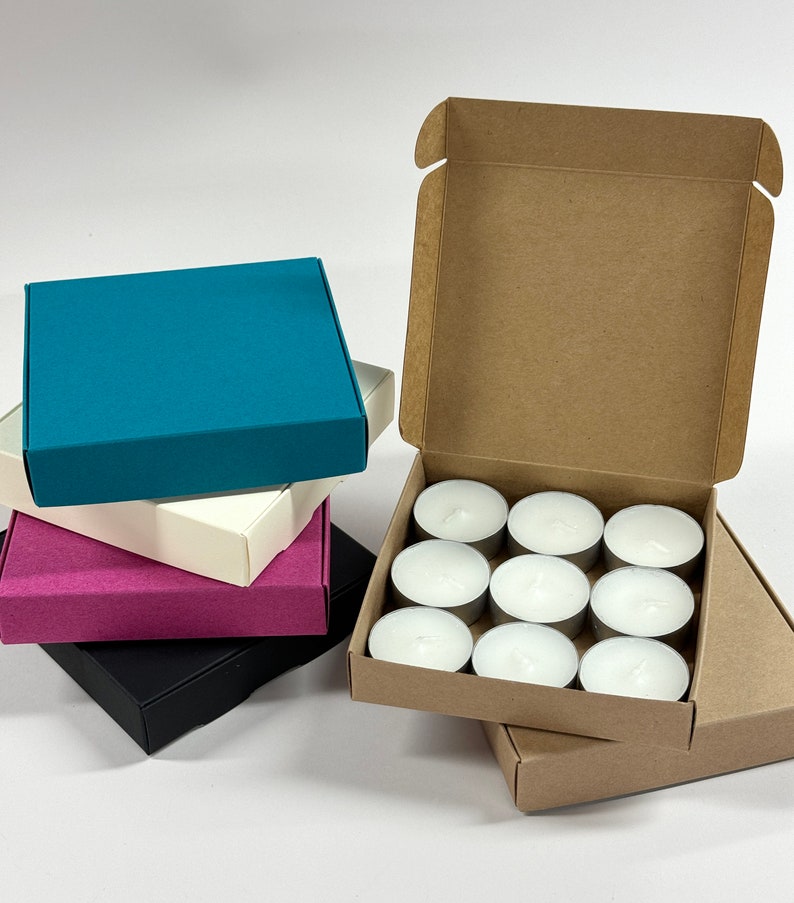 May include: A set of 10 white tea lights in a cardboard box. The box is open and the tea lights are visible. There are four empty boxes stacked on top of the box with tea lights. The boxes are teal, white, pink, and black.