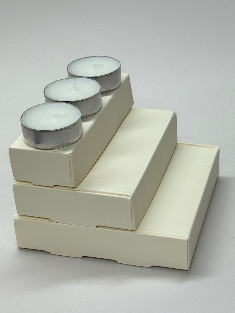 May include: Three white tea lights stacked on top of three white cardboard boxes. The boxes are nested inside each other, creating a tiered effect.