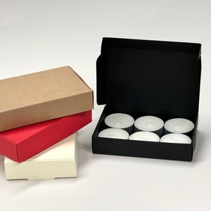 May include: A set of six white tea lights in a black cardboard box. The box is open and the tea lights are visible. There are three boxes stacked on top of each other in the background. The bottom box is white, the middle box is red, and the top box is brown.