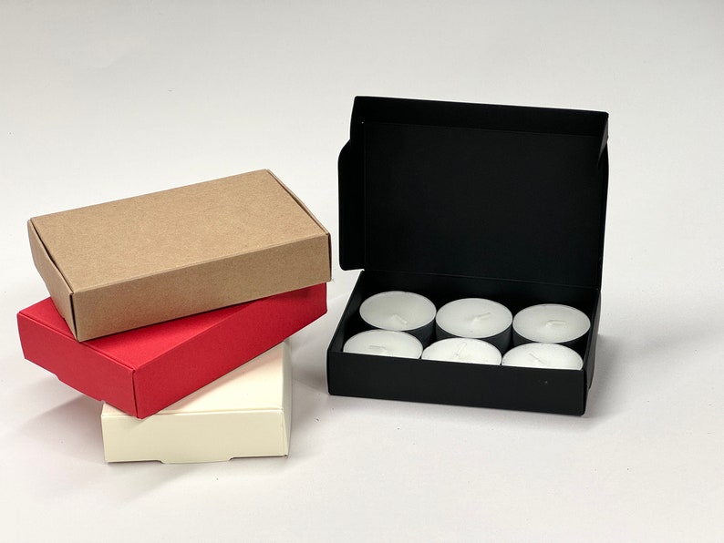 May include: A set of six white tea lights in a black cardboard box. The box is open and the tea lights are visible. There are three boxes stacked on top of each other in the background. The bottom box is white, the middle box is red, and the top box is brown.