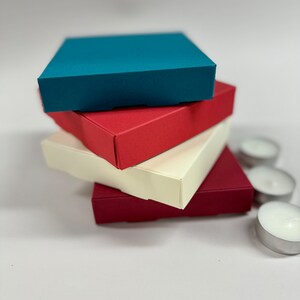 May include: Four square boxes stacked on top of each other. The top box is teal blue, the second box is red, the third box is cream, and the bottom box is burgundy.