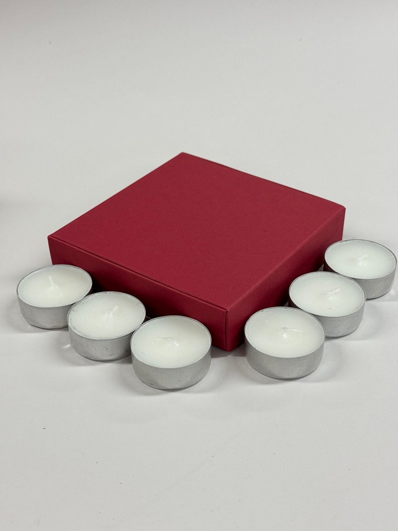 May include: A set of seven white tea lights in silver containers, arranged around a red square box.