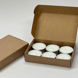 May include: A brown cardboard box with six white tea light candles inside. The box is open and the candles are visible.