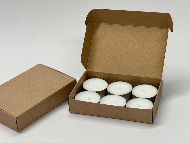 May include: A brown cardboard box with six white tea light candles inside. The box is open and the candles are visible.