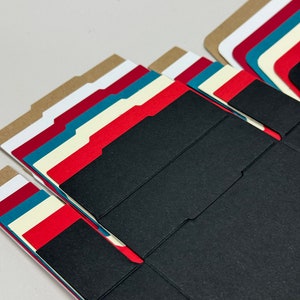 May include: A stack of folded cardboard boxes in various colors, including brown, white, red, teal, and black. The boxes are designed for packaging small items.