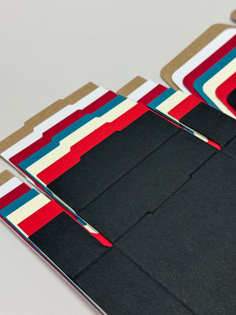 May include: A stack of folded cardboard boxes in various colors, including brown, white, red, teal, and black. The boxes are designed for packaging small items.