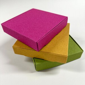 May include: Three square boxes stacked on top of each other. The top box is a bright pink, the middle box is a light brown, and the bottom box is a dark green.