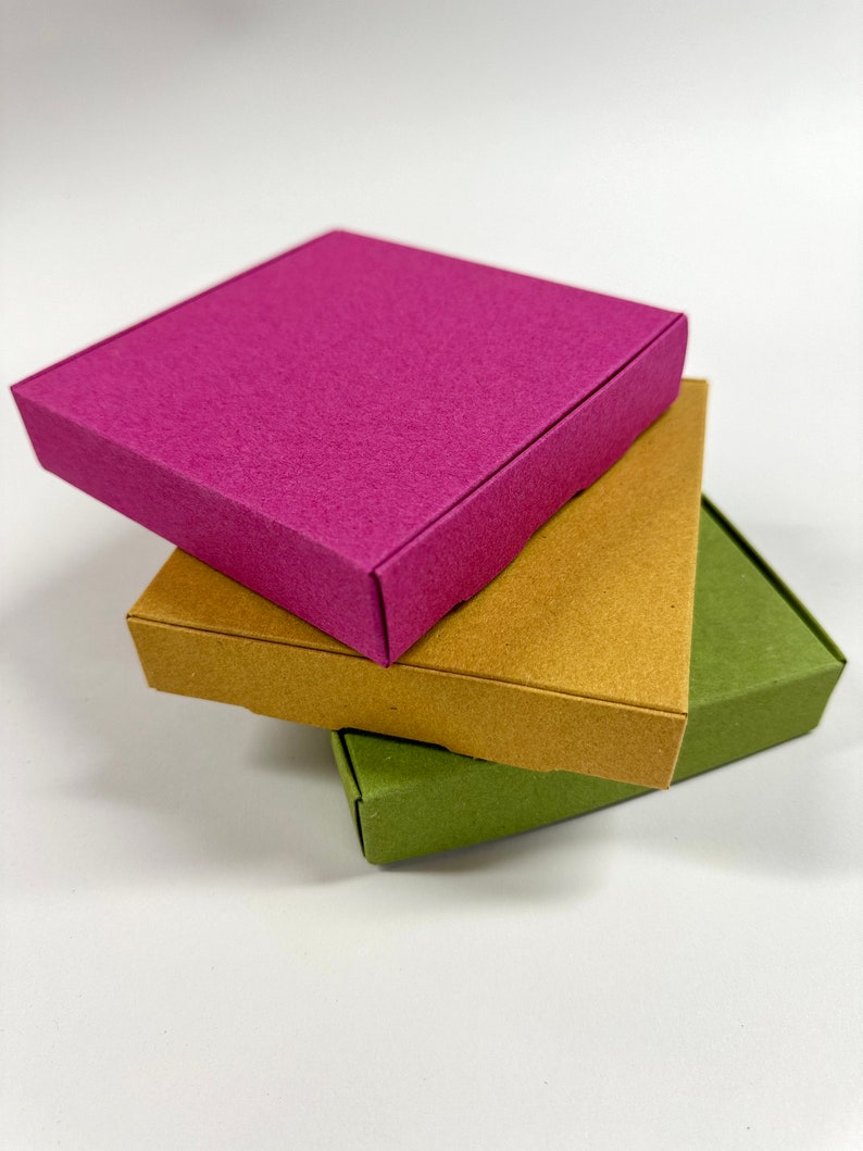 May include: Three square boxes stacked on top of each other. The top box is a bright pink, the middle box is a light brown, and the bottom box is a dark green.