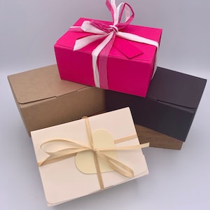 May include: Four gift boxes in different colors and styles. A pink box with a white and pink ribbon, a brown box, a white box with a beige ribbon and a heart-shaped tag, and a black box.