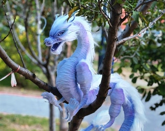 Puede incluir: Un dragón de peluche blanco y morado con una cola larga enroscada alrededor de una rama. El dragón tiene una melena blanca y esponjosa y una cara detallada con una boca abierta.