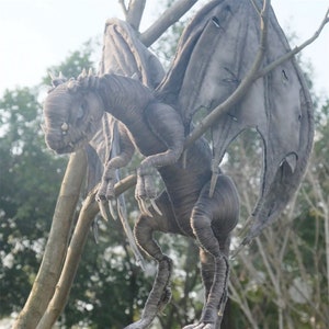 Può includere: Una scultura dettagliata di un drago grigio appollaiato su rami d'albero. Il drago ha la pelle strutturata, grandi ali e artigli affilati. La scultura ha un aspetto realistico, con dettagli intricati e una posa minacciosa. Lo sfondo presenta alberi e fogliame sfocati.