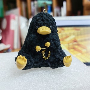 Peut inclure: Un jouet en peluche noir en crochet en forme de Niffleur de la série Harry Potter. Le jouet a une chaîne dorée autour du cou et des pieds jaunes.