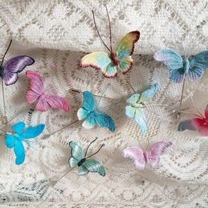 May include: A collection of ten crocheted butterfly pins in various colours, including blue, pink, purple, and green. The pins are displayed on a white lace fabric background.