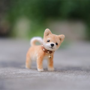 May include: A small, brown and white felt Shiba Inu dog with a brown leather collar and a gold bell. The dog is standing on a gray surface.