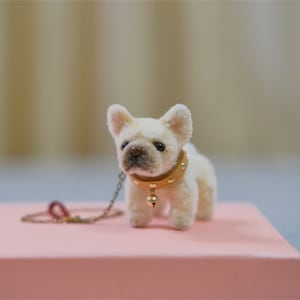 May include: A small, white, plush French bulldog wearing a brown leather collar with gold studs and a gold bell. The dog is on a pink surface and is attached to a gold chain leash.
