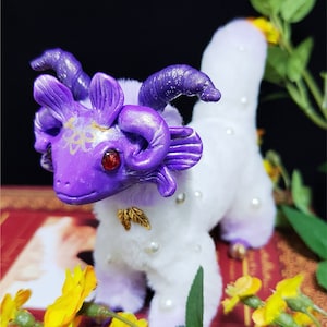 May include: A purple and white furry creature with horns and red eyes. The creature is wearing a pearl necklace and is sitting on a book with yellow flowers.