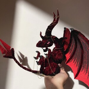 May include: A handcrafted, plush dragon in shades of deep red and black. The dragon features large, detailed wings with a gradient effect, curved horns, and a long tail. Gold-colored chain accents the design, creating a fantasy-themed decorative item.